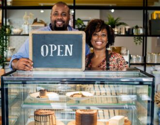 Two local business owners in their newly opened café shop, holding an “Open” sign